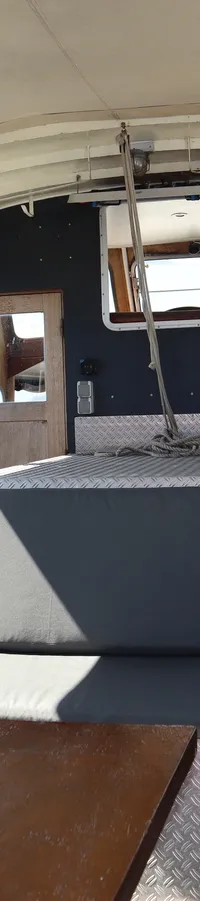 De foto toont het interieur van een boot met een houten tafel, banken, twee deuren en uitzicht op andere boten in een haven.