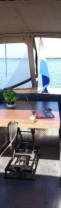 De foto toont de achterkant van een boot met een overkapping, zitbanken, een tafel met laptop en een plant, en een…