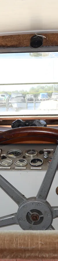 De foto toont het stuurhuis van een boot met een houten stuurwiel, instrumentenpaneel en uitzicht op een haven.