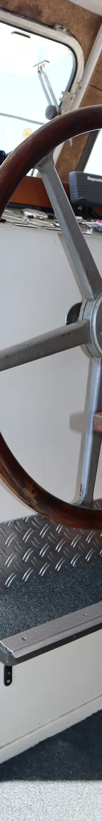 Een houten stuurwiel met metalen spaken, een Raymarine-apparaat en diverse meters en schakelaars zijn zichtbaar.