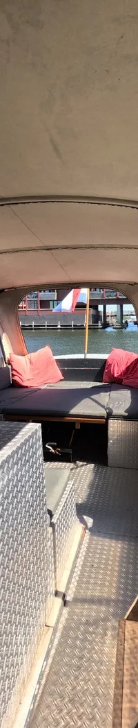 De foto toont het achterdek van een boot met een overkapping, zitbanken met rode kussens en een aluminium opbergkist.