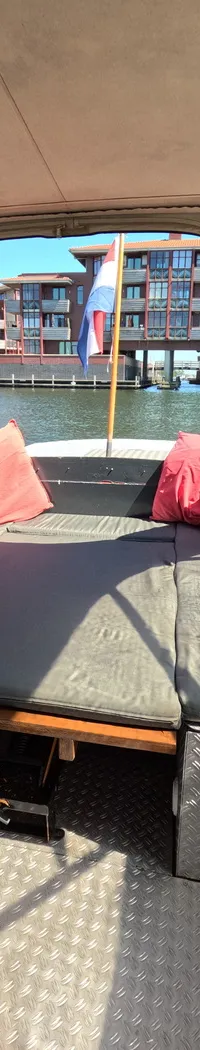 De foto toont het interieur van een boot met een grijze bank, rode kussens, en een Nederlandse vlag aan de achterzijde.