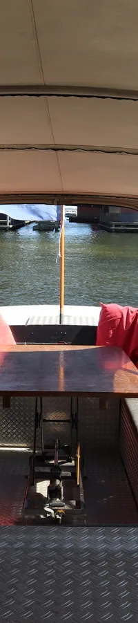 Een overdekte boot met een tafel, banken met kussens en rode sierkussens, en een uitzicht op water en gebouwen.