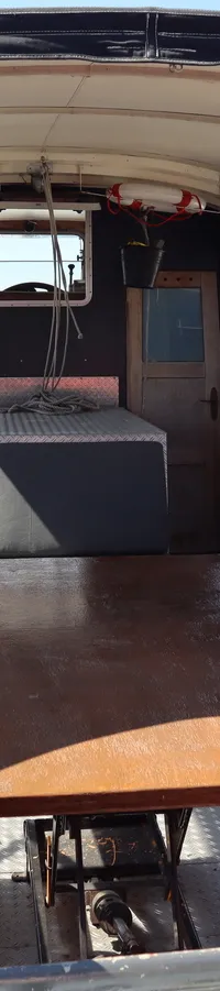 De foto toont het interieur van een boot met een houten tafel, banken met kussens en een afdekzeil met ramen.