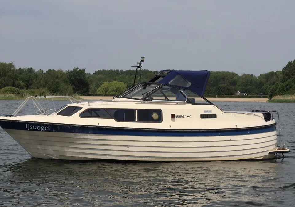 Een witte motorboot met blauwe accenten en de naam "IJsvogel" vaart op een waterweg met bomen op de achtergrond. — Joda 7500