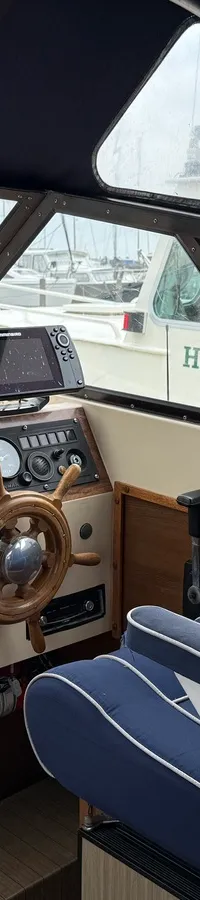 De foto toont de stuurstand van een boot met een houten stuurwiel, diverse meters en schakelaars, en een blauwe stoel.