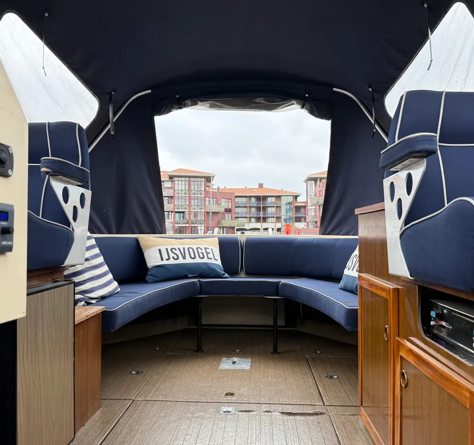 De foto toont het interieur van een boot met blauwe kussens, een kussen met "IJSVOGEL" erop, en houten details. — Joda 7500