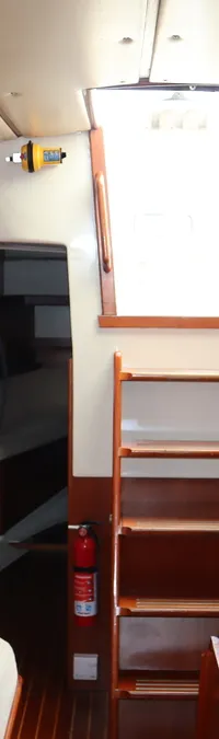 De foto toont het interieur van een bootkajuit met houten lambrisering, witte banken, een kombuis en een trap naar buiten.