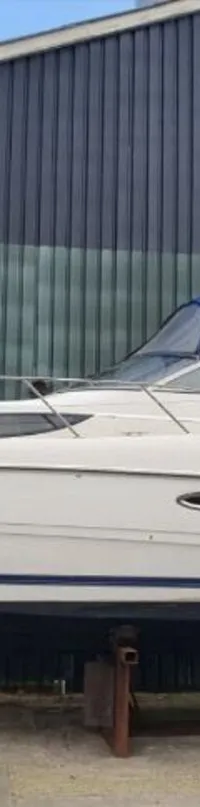 Een witte motorboot met blauwe romp en blauwe cabriokap staat op een bok voor een gebouw. — Bayliner 305 CIERA
