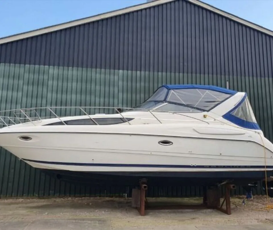Een witte motorboot met blauwe romp en blauwe cabriokap staat op een bok voor een gebouw. — Bayliner 305 CIERA