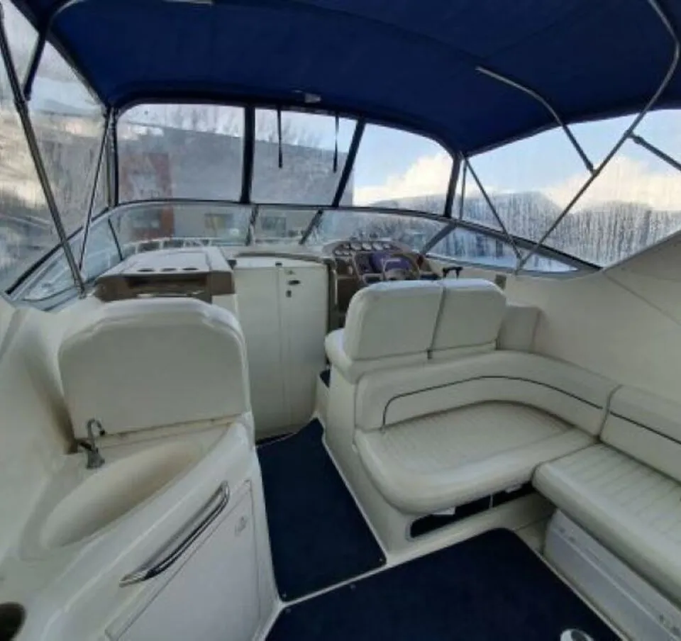 De foto toont het interieur van een boot met een blauwe kap, witte bekleding, een stuurstand, een wasbak en blauwe…