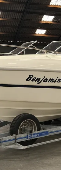 Een witte motorboot met de naam "Benjamin" staat op een dubbelassige trailer in een grote loods. — Cranchi 21 Ellipse