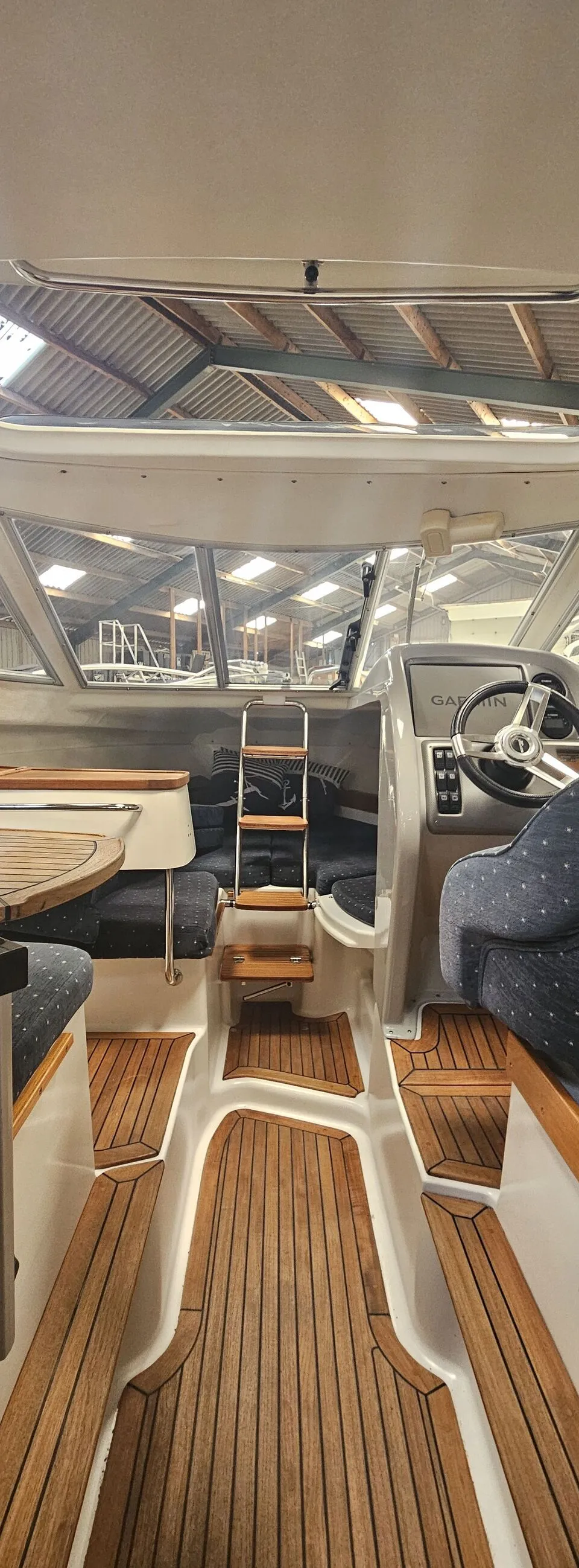 De foto toont het interieur van een boot met een stuurwiel, een Garmin-scherm, houten vloeren en blauwe kussens.