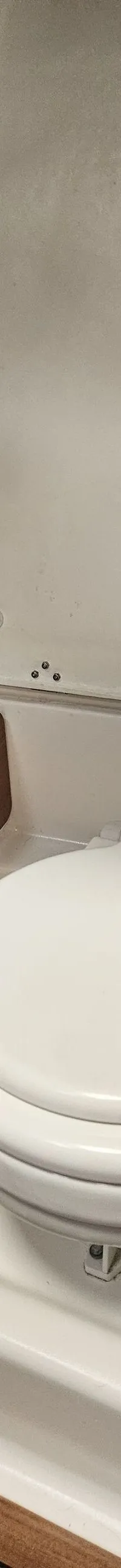 Een witte scheepstoilet met gesloten deksel is zichtbaar, omringd door witte wanden en een houten paneel.