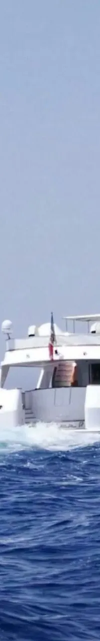 Een witte motorjacht vaart op een blauwe zee, met een vlag aan de achtersteven en een logo van "DE VALK YACHT BROKERS"…