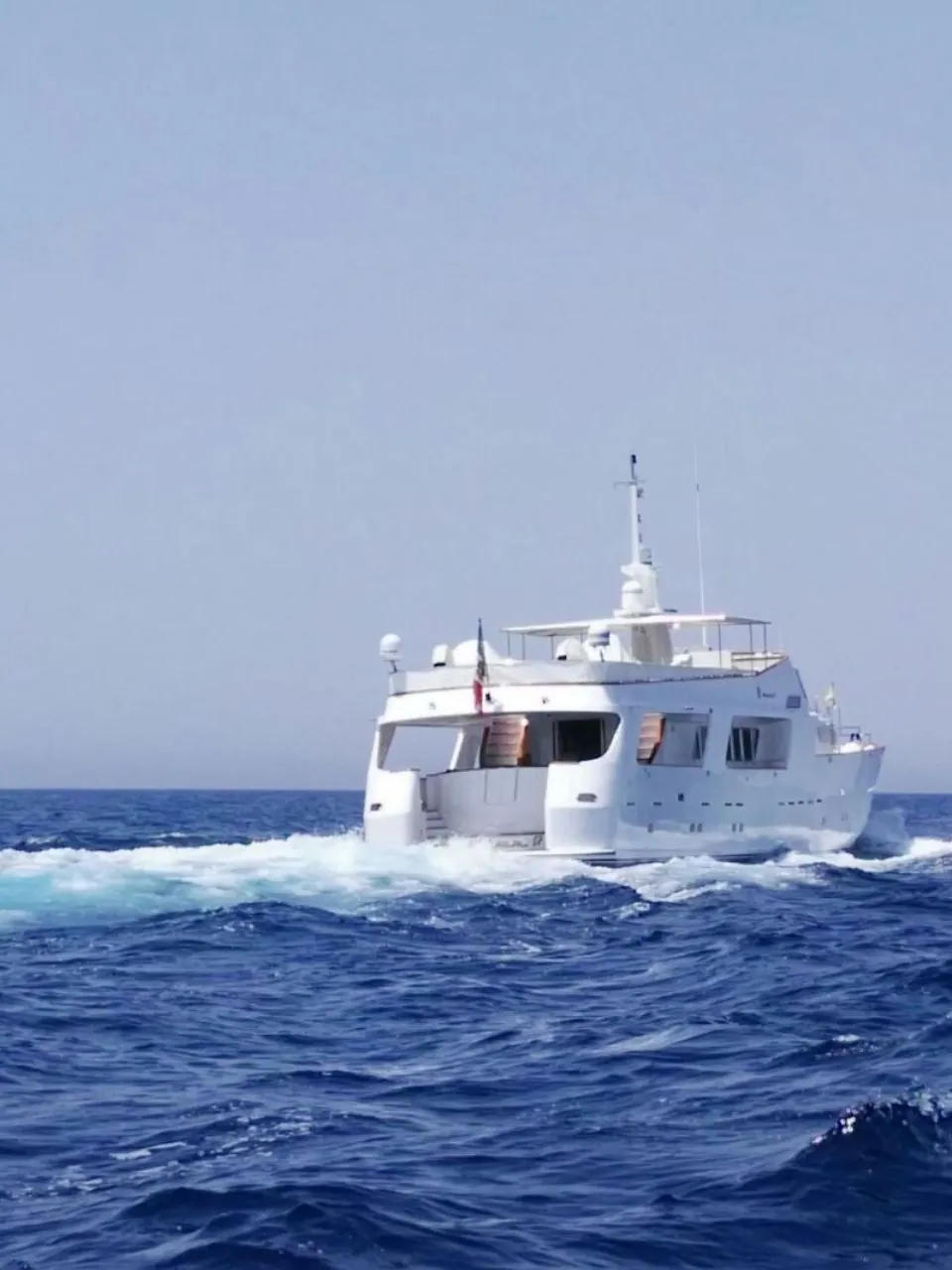 Een witte motorjacht vaart op een blauwe zee, met een vlag aan de achtersteven en een logo van "DE VALK YACHT BROKERS"…