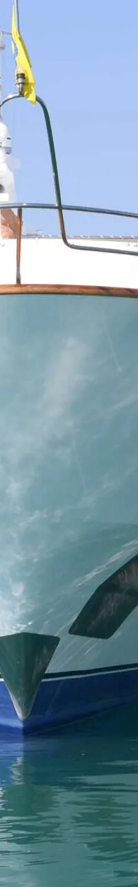 Een lichtblauwe boot met een donkerblauwe onderkant ligt afgemeerd in een haven met een klokkentoren op de achtergrond.