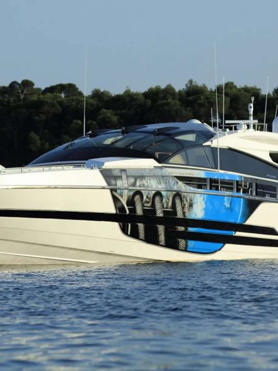 Een witte motorjacht met een blauw-zwart patroon op de romp ligt in het water voor een beboste kustlijn.