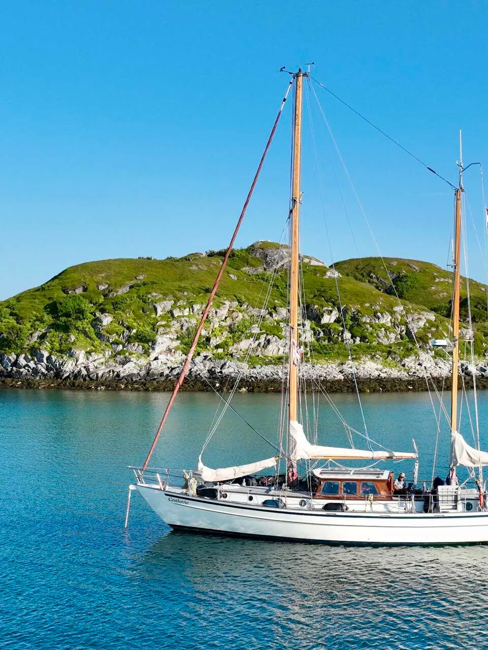 Een witte zeilboot met twee masten ligt op helderblauw water voor een groen, rotsachtig eiland onder een strakblauwe lucht.