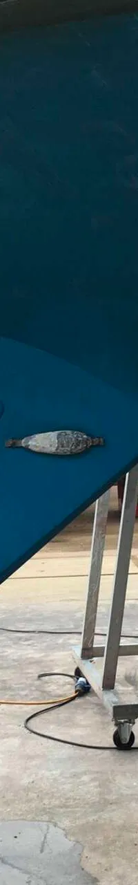 De foto toont de onderkant van een blauwe boot met een roer, schroef en anodes, staand op een trailer in een loods.