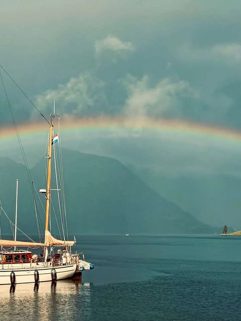 Een witte zeilboot met twee masten en een Nederlandse vlag ligt in een fjord met een regenboog op de achtergrond.