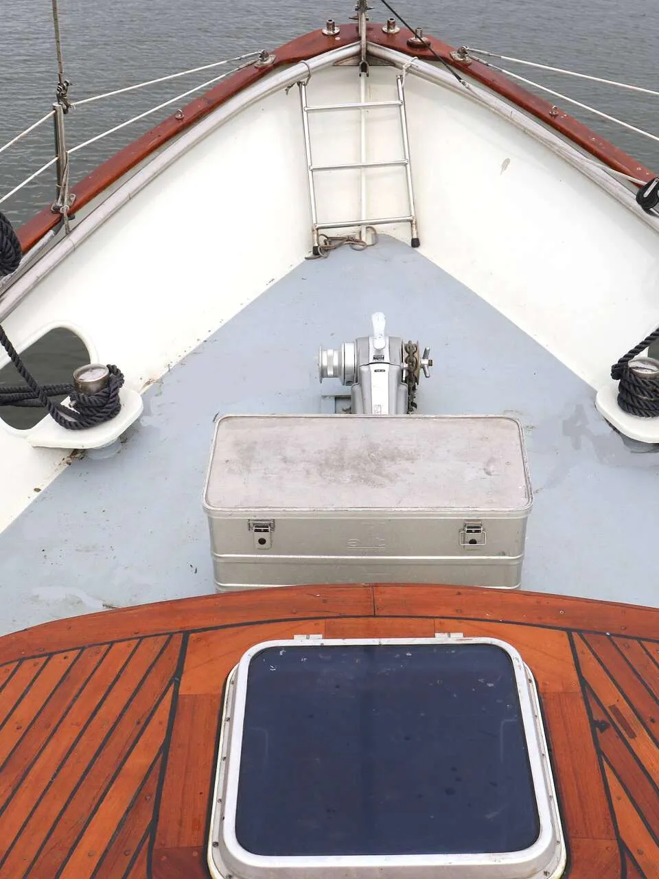 De boeg van een boot is zichtbaar met een houten dek, luik, aluminium kist, ankerlier, touwen en reling.