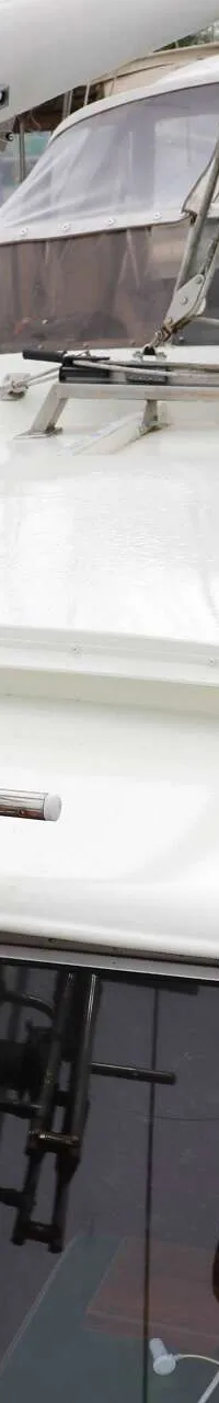 Een witte boot met een mast en een luik op het dek, liggend aan een steiger met andere boten op de achtergrond.