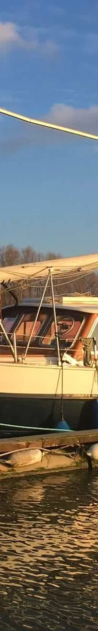 Een schip met een donkere romp en lichte opbouw ligt afgemeerd aan een steiger, met een mast en giek zichtbaar.