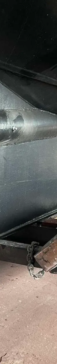 Een close-up van de onderkant van een zwarte boot met een driebladige schroef en een anker, ondersteund door stalen balken.