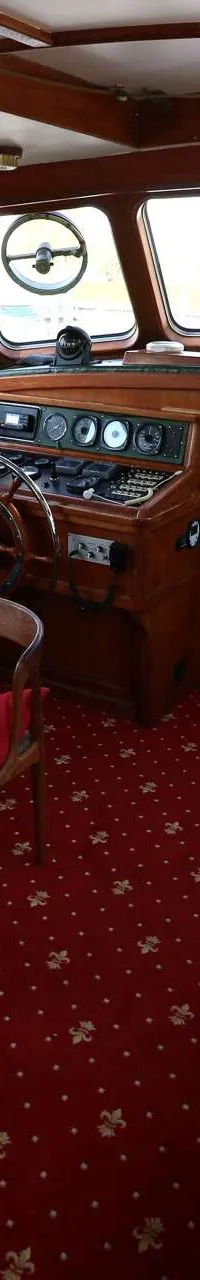 De foto toont het interieur van een boot met een houten stuurstand, een tafel met stoelen, en een rode vloerbedekking.