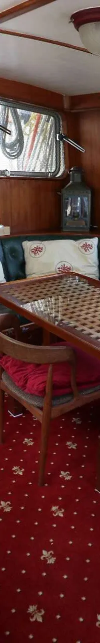 De foto toont een houten interieur van een boot met een rode vloerbedekking, een zithoek, een tafel en een trap.