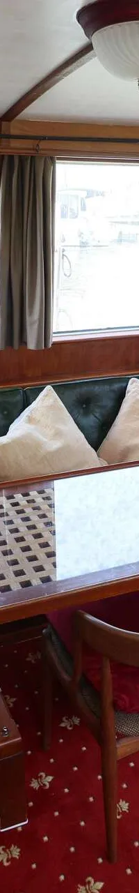 De foto toont een interieur van een boot met een houten stuurstand, een groene leren bank, een tafel en een rood tapijt.