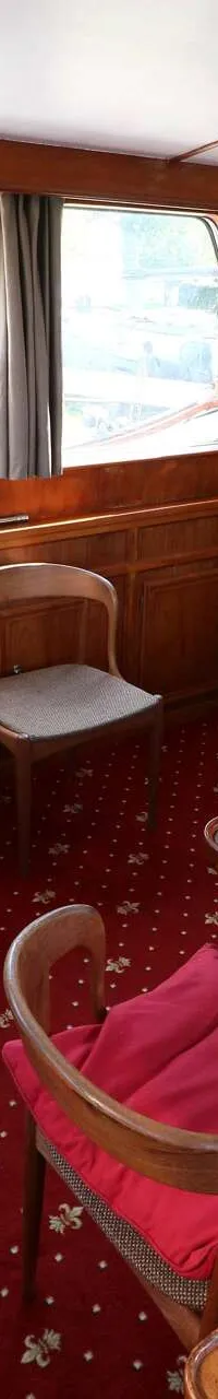 De foto toont een interieur van een boot met houten wanden, een rood tapijt, een tafel, stoelen en een bank.