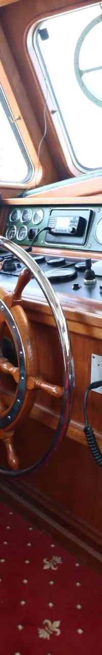 De foto toont een houten stuurhut met een stuurwiel, instrumentenpaneel, radio en een rode vloerbedekking.