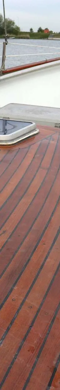 De foto toont het voordek van een boot met een houten dek, een luik, een lier en een reling, met water op de achtergrond.