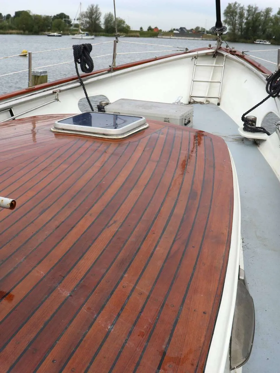 De foto toont het voordek van een boot met een houten dek, een luik, een lier en een reling, met water op de achtergrond.