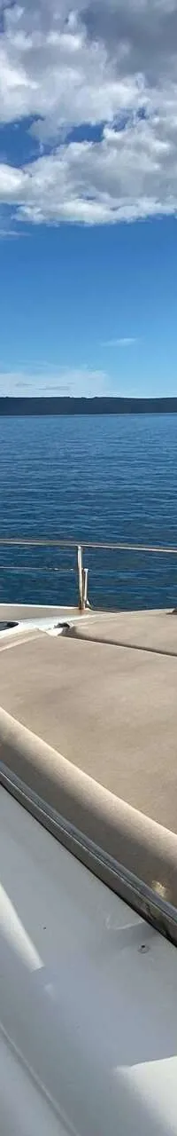 De boeg van een jacht met een zonnedek en houten dek, varend op open water met eilanden op de achtergrond. — Aicon 64