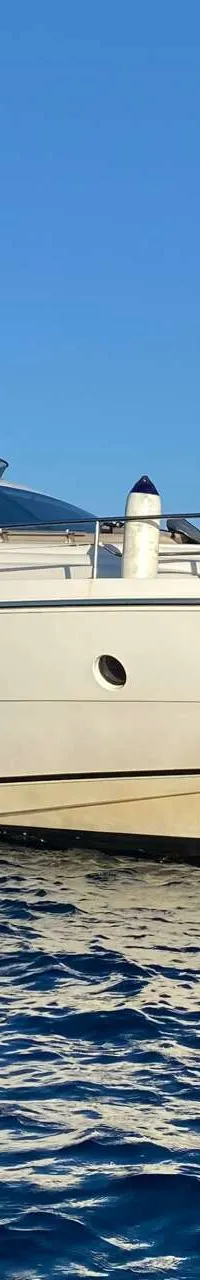 Een witte motorjacht met beige stootwillen en een beige bimini-top ligt op blauw water, met een heuvelachtige kustlijn op…
