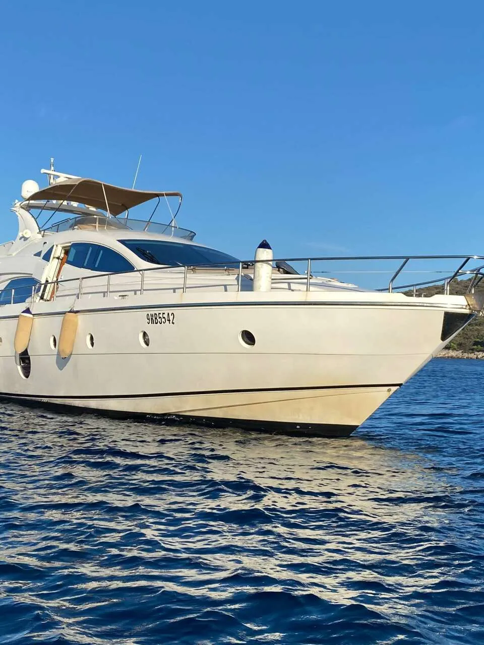 Een witte motorjacht met beige stootwillen en een beige bimini-top ligt op blauw water, met een heuvelachtige kustlijn op…