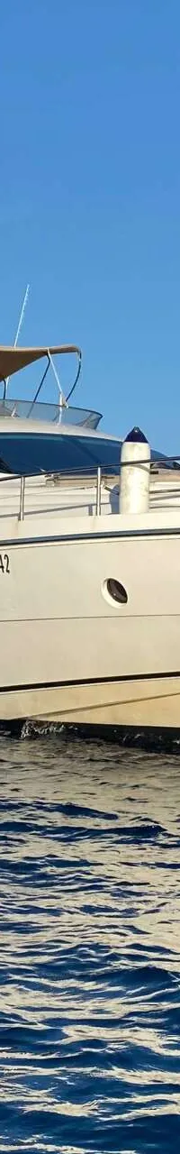 Een witte motorjacht met een bruine bimini-top ligt voor anker in een blauwe baai met rotsachtige oevers.