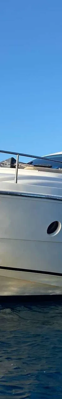 Een witte motorboot met een beige zonnescherm ligt voor anker in blauw water, met rotsachtige kustlijn op de achtergrond.