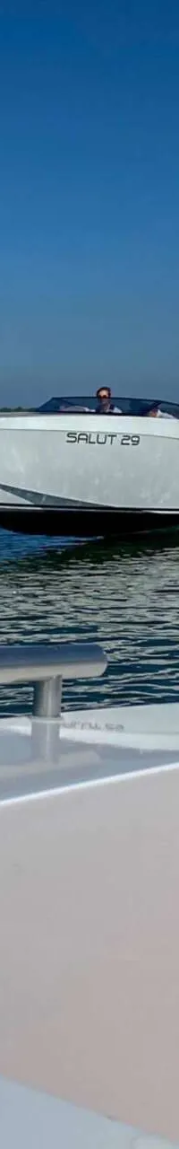 Een witte boot met opschrift "SALUT 29" vaart snel over water, gezien vanaf een andere boot, met een blauwe lucht en…