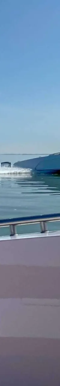 Een lichtgekleurde boot vaart snel op rustig water, gezien vanaf een andere boot met een railing en een bekerhouder.
