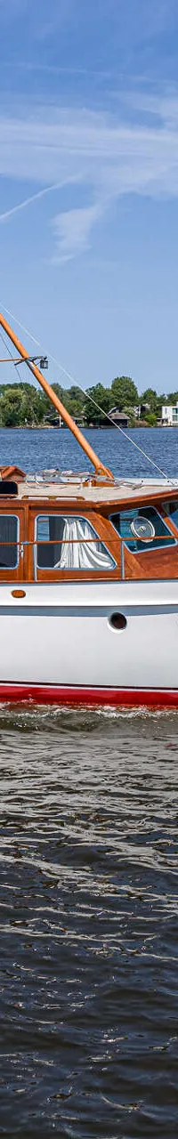 Een witte motorboot met houten opbouw en een rode waterlijn vaart over rustig water onder een blauwe lucht.