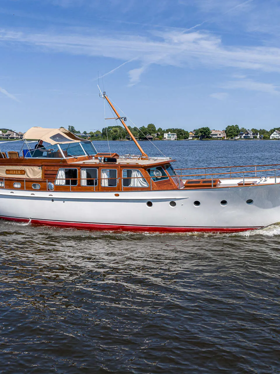 Een witte motorboot met houten opbouw en een rode waterlijn vaart over rustig water onder een blauwe lucht.