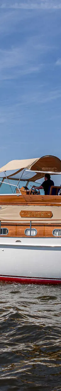 Een witte motorboot met houten opbouw en een rode onderkant vaart op het water onder een blauwe lucht.