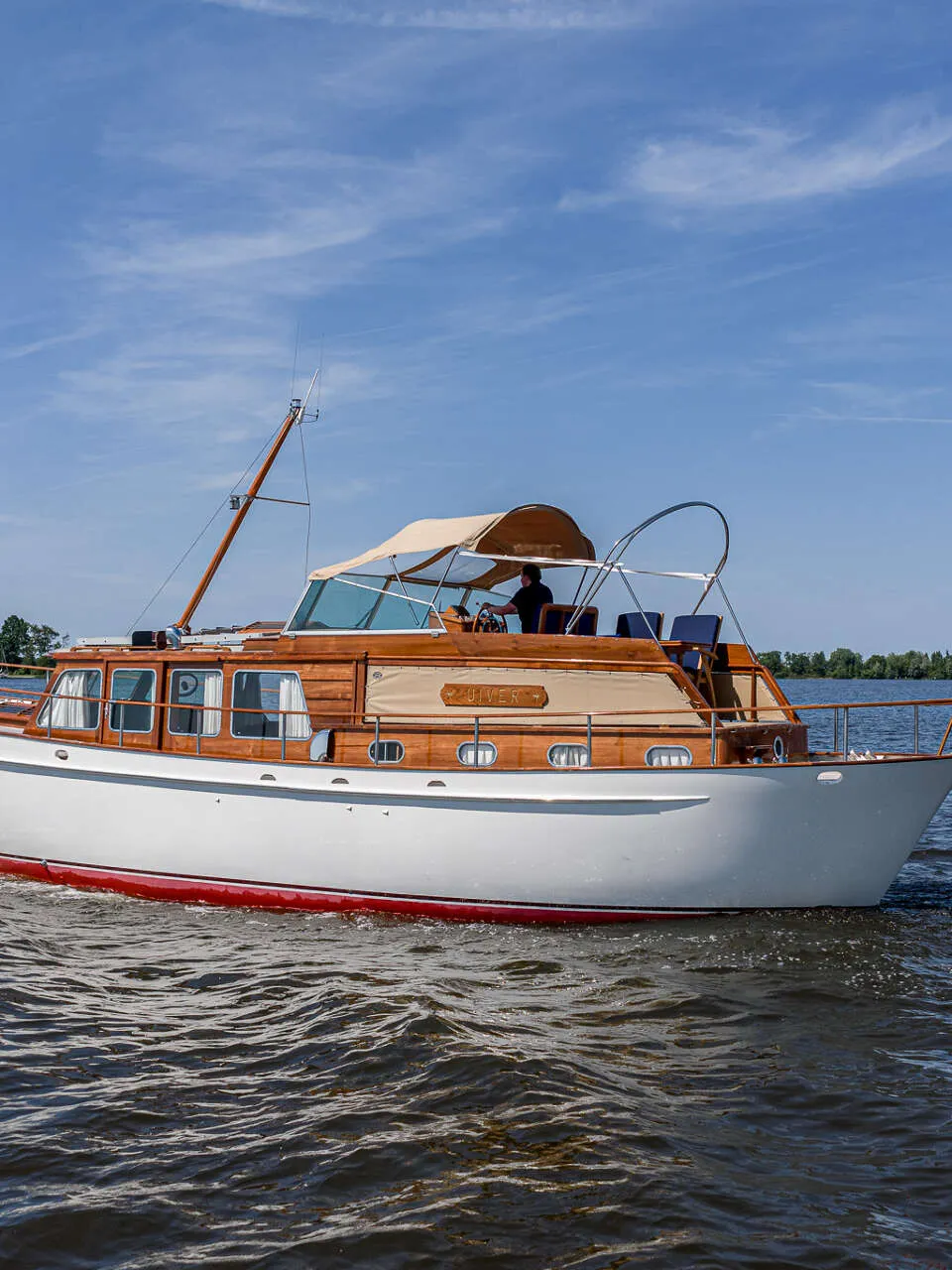 Een witte motorboot met houten opbouw en een rode onderkant vaart op het water onder een blauwe lucht.
