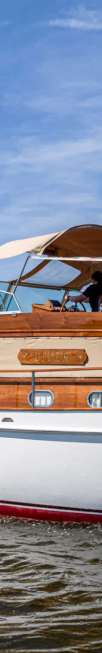 Een motorjacht met een witte romp, rode onderkant en houten opbouw vaart op een waterweg onder een blauwe lucht.