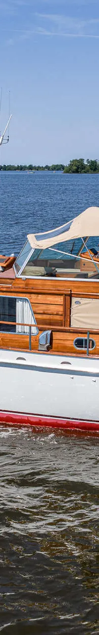 Een witte motorboot met houten opbouw en een rode streep op de romp vaart op een meer onder een blauwe lucht.