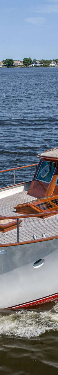 Een witte motorboot met houten opbouw vaart op een meer met huizen aan de oever onder een blauwe lucht.